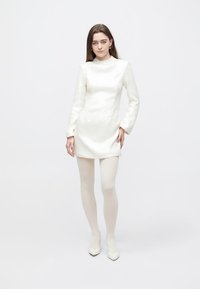 Biele mini šaty s dlhými rukávmi, textúrovanou látkou a vysokým golierom. Nosené s bielymi pančuchami a lodičkami s špicatou špičkou. Jednoduchý dizajn.