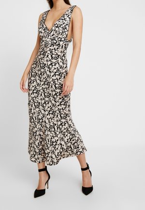 Kvinde iført en ærmeløs sort og hvid blomsterprint midi-kjole med dyb V-udskæring og sideskæringer, kombineret med sorte ankelrem-sandaler.