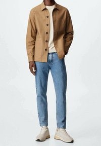 Veste marron à boutons avec un col et des poches avant, portée sur une chemise claire, associée à un jean bleu clair et des baskets beiges.