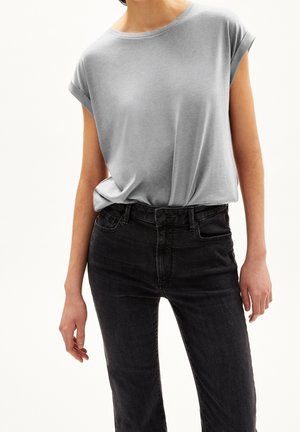 Osoba ubrana w jasnoszarą, włożoną do środka koszulę z krótkim rękawem oraz czarne, wysokie spodnie dżinsowe na tle jednolitego jasnego tła.