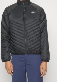 Černá péřová bunda se zipem vpředu, vysokým límcem a gumovým logem Nike. Má prošívaný vzor a elastické manžety a spodní lem.