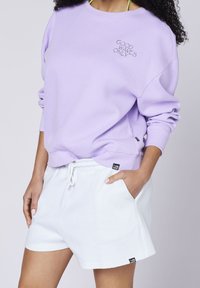 Sudadera de lavanda con textura acanalada y bordado "SOLO BUENAS OLAS". Combinada con shorts blancos de corte holgado con bolsillos laterales.