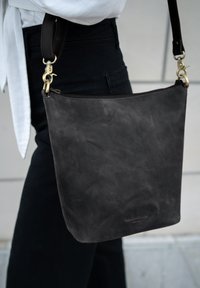 Schwarze Lederhandtasche mit glatter Textur, Reißverschluss und verstellbarem Schulterriemen mit Messingclip. Minimale Prägung des Logos an der Seite.