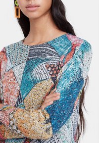 Maglione lavorato a maglia con design patchwork caratterizzato da motivi multicolori in blu, arancione e nero. Texture morbida con grafiche variegate e accenti scintillanti.