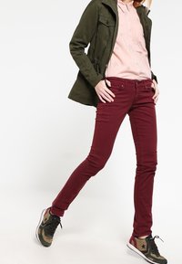Jean skinny bordeaux foncé associés à une chemise rose clair avec des motifs d'étoiles et une veste verte olive. Baskets avec accents camouflés.