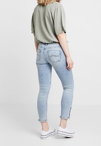 Jeans skinny azzurri con una vestibilità a vita media, dotati di tasche posteriori e cerniere laterali sulle caviglie, abbinati a una blusa larga verde oliva.
