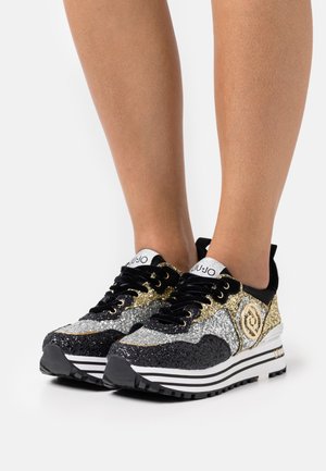 Legs wearing black, silver, and gold glittery sneakers with black laces and striped white and black soles, posed against a plain white background.