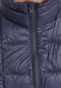 Mornarska puffer jakna s visokim ovratnikom, punom dužinom zatvarača i kontrastnim šavovima, s glatkom, sjajnom teksturom.