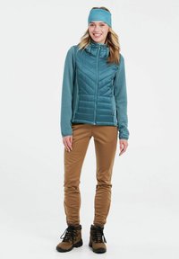 Mėlyna šilta liemenė su tekstūruotomis rankovėmis, rudos prilipusios kelnės ir tamsūs žygio batai. Plaukų juosta derinama su liemenės spalva.