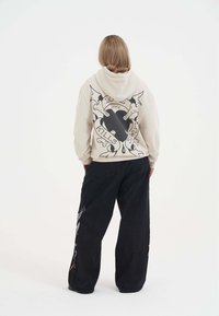 Beżowa bluza z kapturem z czarnym grafikiem przedstawiającym serce, sztylety i tekst na plecach. Czarne szerokie spodnie z kolorowymi detalami po bokach.