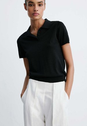 Femme portant un polo noir à manches courtes rentré dans un pantalon blanc plissé taille haute, les mains dans les poches.