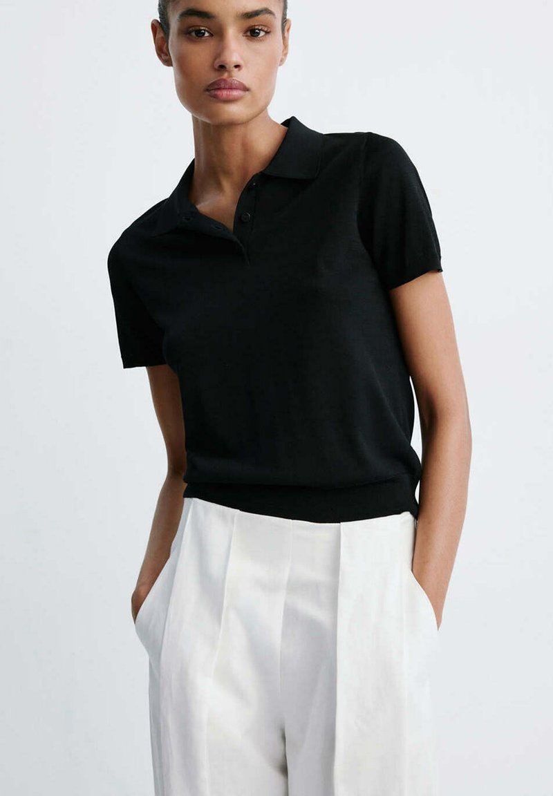 Femme portant un polo noir à manches courtes rentré dans un pantalon blanc plissé taille haute, les mains dans les poches.