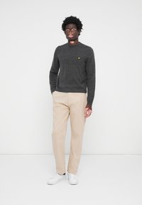 Pull en tricot gris avec un col rond, logo jaune sur la poitrine, associé à un pantalon beige et des baskets blanches. Texture lisse et coupe ajustée.