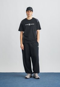 Μαύρο t-shirt με κοντά μανίκια της Nike και λογότυπο, συνδυασμένο με φαρδιά μαύρη παντελόνια και γκρι αθλητικά παπούτσια. Το μοντέλο φορά επίσης μαύρο καπέλο. Απλός, αθλητικός στυλ.
