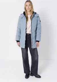 Ljusblå puffjacka med diagonalt quiltning, dragkedja fram och avtagbar huva. Buren över en ljus tröja och svarta jeans.