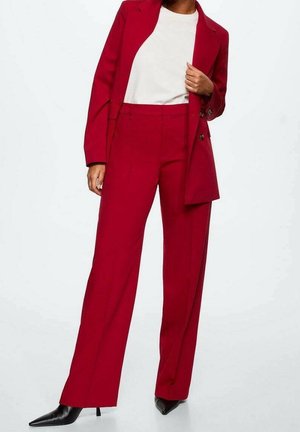 Frau trägt ein maßgeschneidertes rotes Blazer und dazu passende, weit geschnittene Hosen, kombiniert mit einem weißen Oberteil und schwarzen Schuhen mit spitzer Spitze.