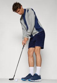 Grå og marineblå genser, korte marineblå corduroy-shorts, marineblå golfsko med blå aksenter, stående, forbereder seg til å svinge golfkølle ved siden av en ball.
