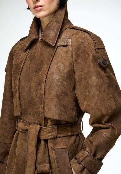 Žena v hnedom semišovom trenírkovom plášti so širokým golierom, opaskom v páse, náprsnými dierkami a klopovými vreckami na bielom pozadí.