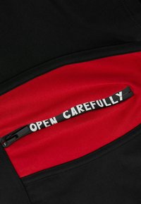 Czarna i czerwona tkanina z kieszenią na zamek, wyposażoną w biały uchwyt z napisem „OTWIERAJ OSTROŻNIE”, kontrastujące faktury i kolory.