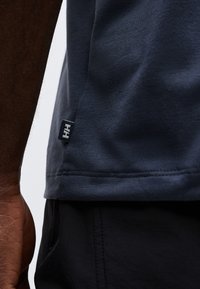 Κοντινό πλάνο ενός προσώπου που φοράει σκουρό μπλε πουκάμισο με μικρή ετικέτα "HH" κοντά στο τελείωμα και μαύρο παντελόνι, με ορατό μέρος του δεξιού του χεριού.