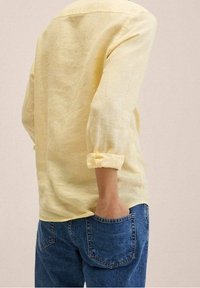 Chemise en lin jaune clair à manches retroussées, associée à un jean en denim bleu. La chemise présente une coupe décontractée et des détails de couture simples.