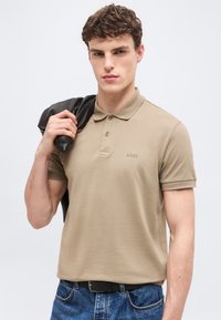 Νέος άνδρας που φοράει μπεζ πουκάμισο πόλο και μπλε τζιν, κρατώντας ένα μαύρο σακάκι στον ώμο του, στέκεται μπροστά σε απλό λευκό φόντο.