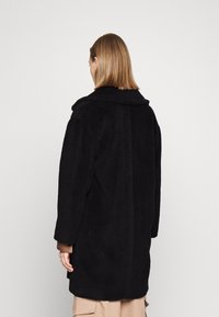 Zwarte bontjas met een kraag, lange mouwen en rechte snit. Textuur aan de oppervlakte en tot onder de heupen. Model is van achteren bekeken.