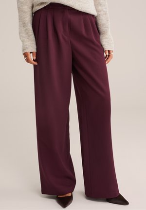 Mujer con pantalones morados oscuros de talle alto y pierna ancha con pliegues, jersey beige, anillos en los dedos y zapatos negros puntiagudos.
