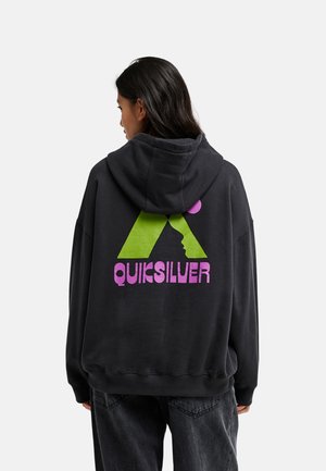 Personne aux longs cheveux foncés portant un sweat à capuche noir avec un logo Quiksilver vert et violet dans le dos, debout sur un fond uni.