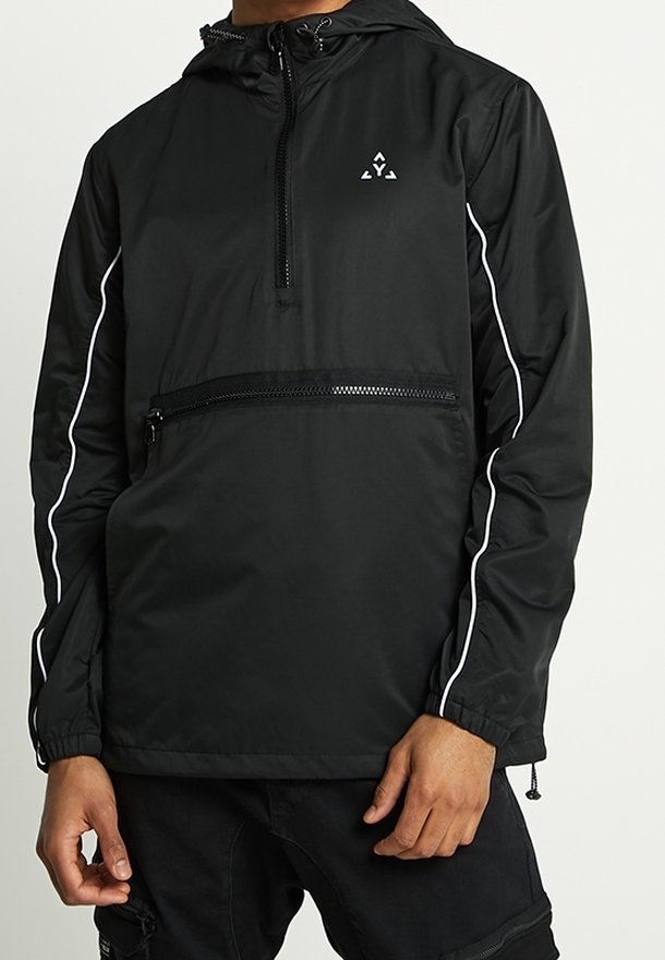 Coupe-vent noir à capuche avec poche zippée devant, passepoil blanc le long des manches et petit logo blanc sur la poitrine, porté par une personne avec les mains visibles.