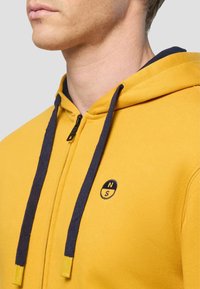 Primo piano di una persona che indossa una felpa gialla con zip, con cordini neri e un piccolo logo circolare "N S" sul petto.