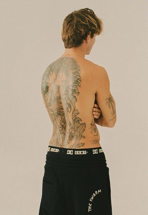 Zwarte shorts met "THE SWARM" in witte tekst, gedragen door een persoon met zichtbare tatoeages op de rug en armen, lichte achtergrond.