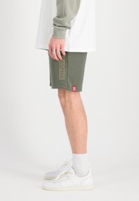 Shorts en coton vert olive avec un logo imprimé "ALPHA", associés à des baskets et des chaussettes blanches. Texture lisse, coupe à la longueur des genoux.