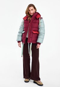 Nő bordó és világoskék kapucnis steppelt kabátban, bő szárú barna nadrágban és barna túrabakancsban, fehér háttér előtt állva.
