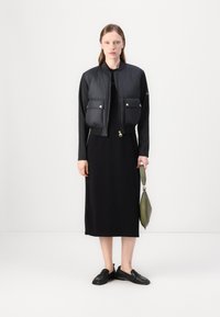 Μαύρο ραμμένο μπομπερ τζάκετ με χρυσές λεπ�τομέρειες, φορεμένο πάνω από ένα μαύρο φόρεμα. Το μοντέλο κρατάει μια ελιά πράσινη τσάντα και φοράει μαύρα slip-on παπούτσια.