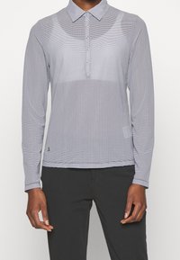 En person klädd i en långärmad, ljusgrå mönstrad pikétröja och svarta byxor, som står framför en vit bakgrund.