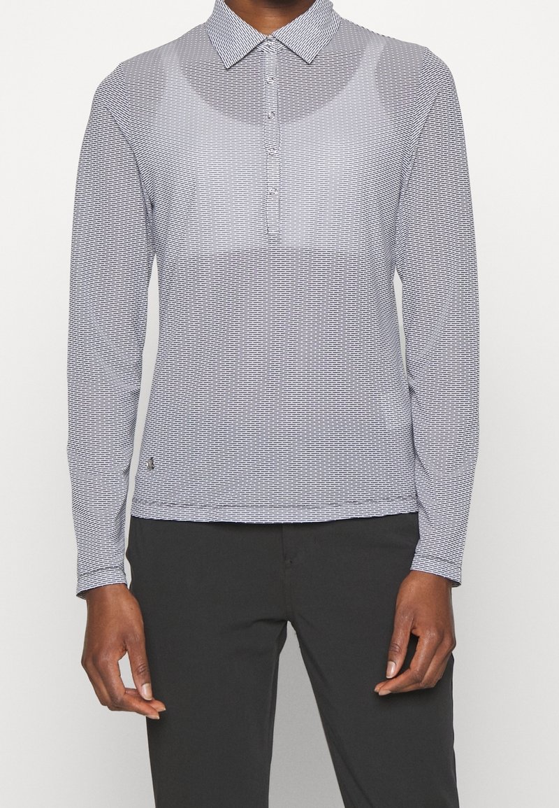 En person klädd i en långärmad, ljusgrå mönstrad pikétröja och svarta byxor, som står framför en vit bakgrund.