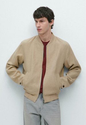 Giovane uomo con capelli scuri che indossa una giacca beige con cerniera sopra una maglietta rossa e jeans grigi chiari, in piedi davanti a uno sfondo chiaro e semplice.