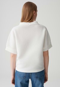 Blusa bianca con texture, maniche corte e colletto pieghettato, abbinata a jeans in denim blu. Design semplice e minimalista con una finitura pulita.