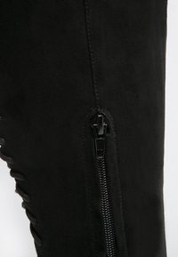 Stivale sopra il ginocchio in camoscio nero con cerniera laterale. Presenta una trama liscia e un laccio decorativo lungo la cucitura esterna.