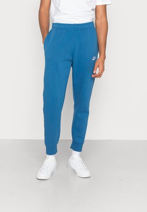 Person trägt blaue Nike-Sweatpants, weiße Socken, weiße Sneaker und ein weißes T-Shirt und steht vor einem einfarbigen hellen Hintergrund.