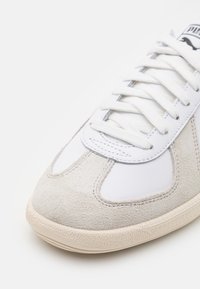 Weißer Sportschuh mit Obermaterial aus Wildleder und Leder, flachen weißen Schnürsenkeln, gepolsterter Zunge und strukturiertem Gummiaußensohle. Mit einem Logo auf der Zunge.