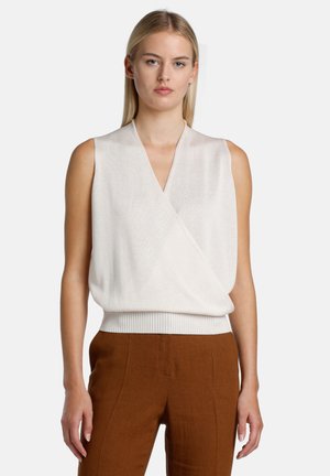 Donna che indossa un top avvolgente smanicato color avorio e pantaloni marroni a vita alta, in piedi contro uno sfondo chiaro e uniforme.