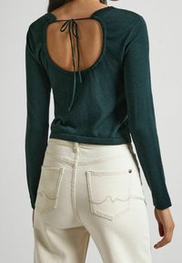 Długi rękaw ciemnozielonego topu z okrągłym otworem na plecach, związanym pasującą wstążką, w połączeniu z jasnokremowymi dżinsami ozdobionymi przeszyciami w kształcie fal.