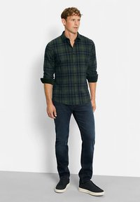 Grün und marineblau karierte Hemd mit langen Ärmeln, kombiniert mit dunkler Jeans und schwarzen Sneakers. Das Hemd hat eine schlanke Passform und Knöpfe an den Manschetten.