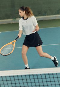 Atleetlik varustus valge lühikesekujulise t-särgi ja musta tennise seelikuga. Hoides oranži tennisereketti, sinisel siseväljakul võrgu ees.