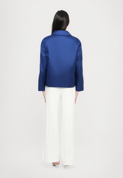 Blå satengjakke med spiss krage, lange ermer og rett skjæring, iført over hvite plisserte bukser.