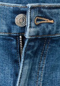 Gros plan sur un jean en denim bleu avec un bouton métallique visible, des coutures marron et une fermeture éclair partiellement ouverte.