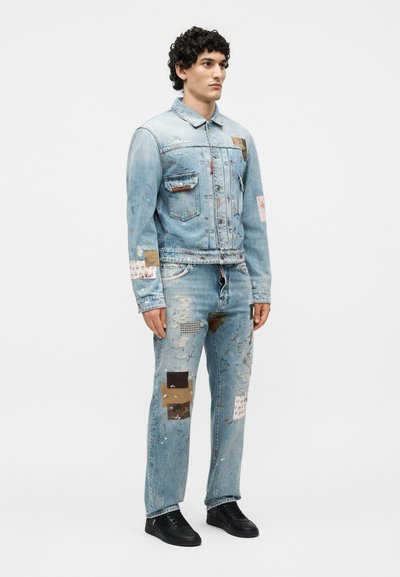 Άνδρας που φορά ανοιχτό μπλε τζιν μπουφάν και τζιν με διάφορα υφασμάτινα patches και πιτσιλιές από μπογιά, φοράει μαύρα αθλητικά παπούτσια.
