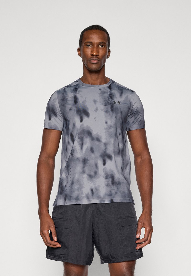 Homme debout de face, portant un t-shirt de sport tie-dye gris et noir et un short noir sur un fond uni.